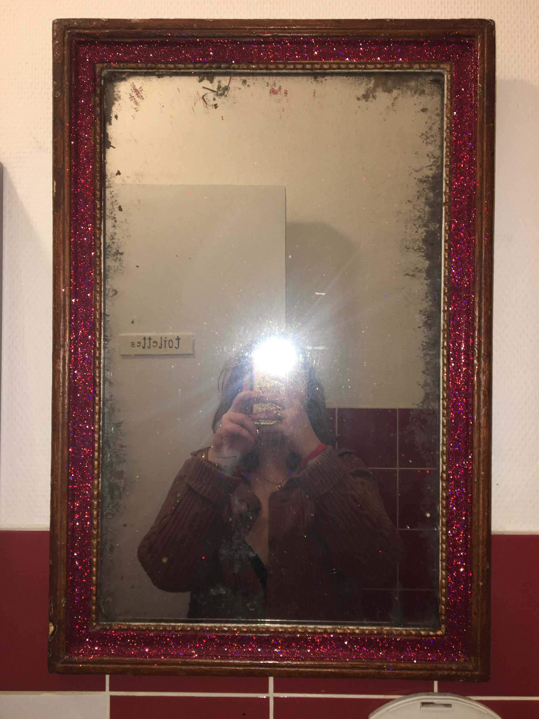 Un miroir assez ancien est installé dans des toilettes. Le cadre en bois a été recouverte de peillettes rouges et quelqu'un se prends en photo en selfie dedans.